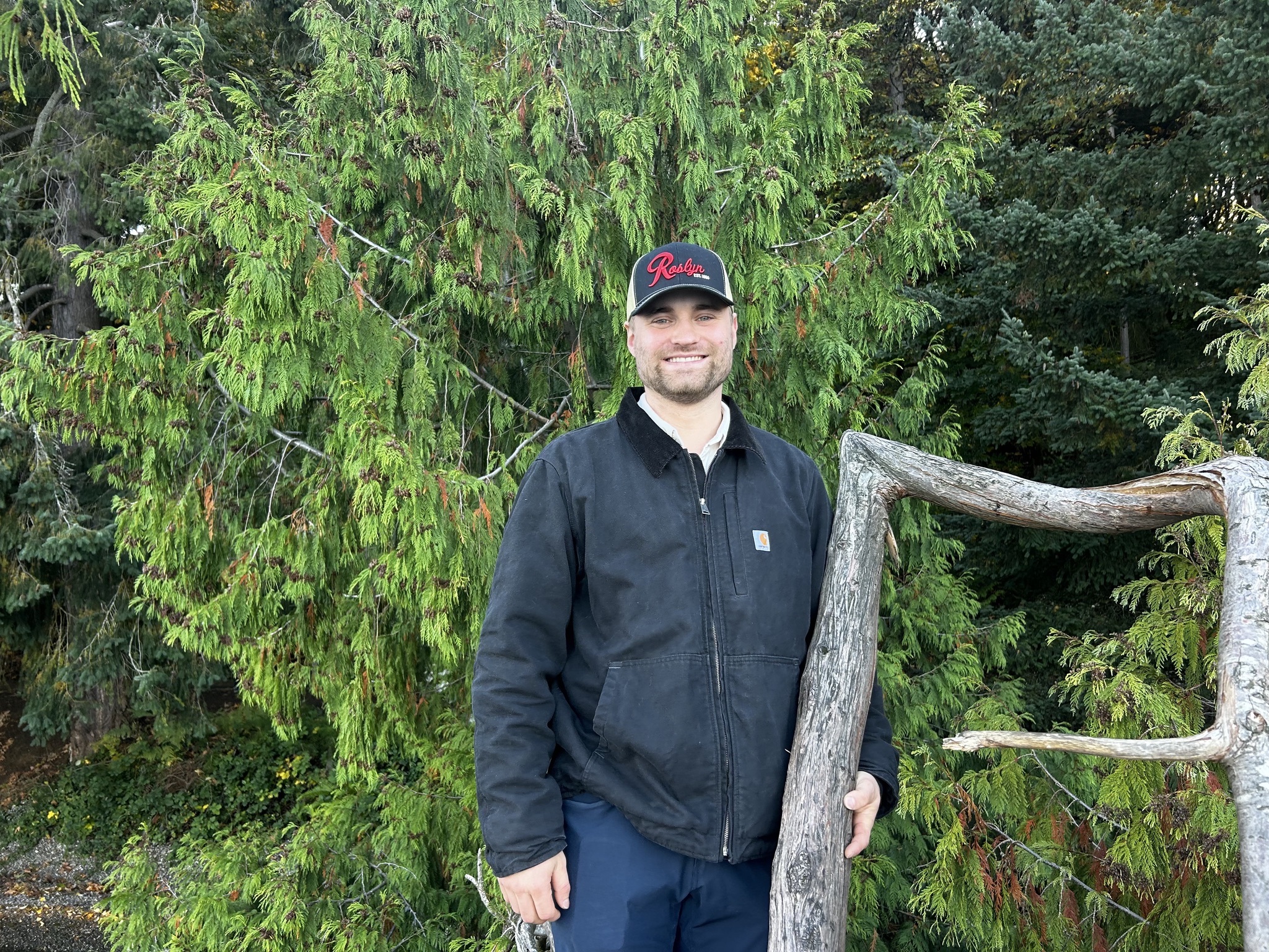 Andy, partner and carpenter, standing outdoors in front of evergreens